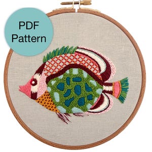 Puede incluir: Diseño de pez bordado en un aro de madera. El pez presenta un patrón colorido con tonos de rosa, verde, rojo y amarillo. El texto "PDF Pattern" está en un círculo azul en la parte superior.