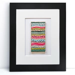 May include: A framed artwork featuring a rectangular textile piece with colorful horizontal bands. The bands include pink, green, blue, and yellow, with embroidered patterns. The frame is black with a white mat.