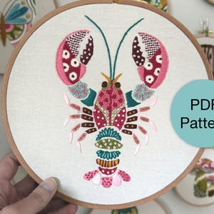 May include: Embroidered lobster design in pink, red, teal, and yellow thread on a cream-colored fabric, set in a wooden embroidery hoop. The design is a PDF pattern. Other embroidery hoops with designs are visible in the background.
