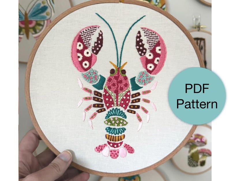 May include: Embroidered lobster design in pink, red, teal, and yellow thread on a cream-colored fabric, set in a wooden embroidery hoop. The design is a PDF pattern. Other embroidery hoops with designs are visible in the background.