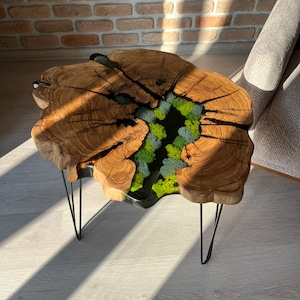 May include: A wooden coffee table with a live edge top and a black epoxy resin fill. The resin fill features a green moss design. The table has black hairpin legs.