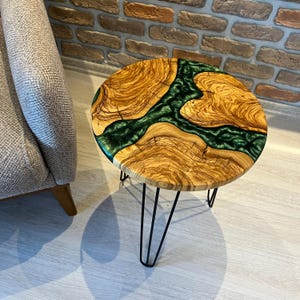 May include: Round wooden side table with a green epoxy resin inlay. The table has a natural wood finish and black hairpin legs.
