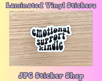 Könnte beinhalten: Ein schwarz-weißer Aufkleber mit dem Text "emotional support kindle" in einer Retro-Schriftart. Der Aufkleber befindet sich auf einem Holzgrund.
