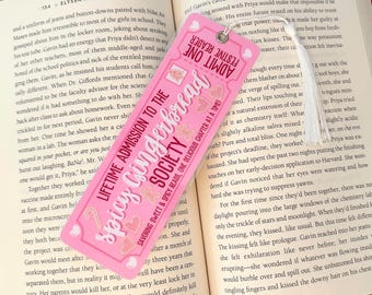 Puede incluir: Marcapáginas rosa con temática de pan de jengibre. El marcapáginas tiene un diseño de boleto con el texto "Admisión de por vida a la Sociedad de Pan de Jengibre Picante" y "Disfrutando de lecturas dulces y picantes, un capítulo delicioso a la vez!"