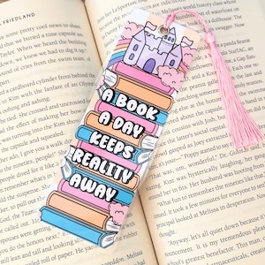 May include: A colorful bookmark with a stack of books and a castle on top. The bookmark has the text "A Book A Day Keeps Reality Away".