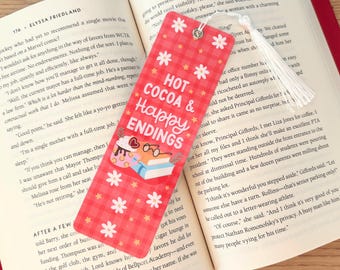 May include: A red bookmark with the text "HOT COCOA & Happy ENDINGS" and illustrations of a cupcake, books, and glasses. The bookmark has a white tassel and is placed in an open book.