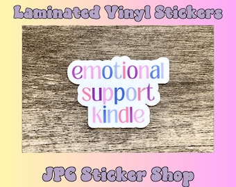 May include: A white sticker with the words "emotional support kindle" in pink, blue, and purple letters on a brown wooden background. The sticker is for sale in a shop called "JPC Sticker Shop".
