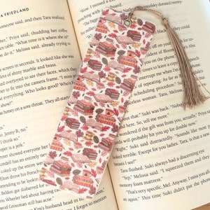 May include: A bookmark with a white background and a pattern of stacked books, leaves, and coffee cups in shades of pink, brown, and orange. The bookmark has a tassel.