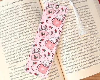 May include: Pink and white bookmark with a repeating pattern of mugs of hot chocolate with whipped cream and candy canes. The bookmark has a white tassel.