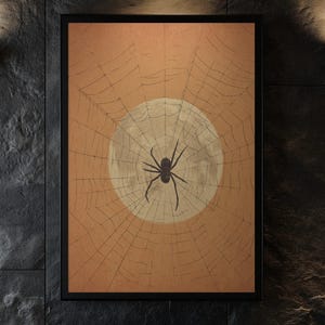 May include: Framed artwork featuring a spider in a web against a full moon backdrop. The spider is black, the web is light brown, and the moon is white. The artwork is framed in a black rectangular frame.