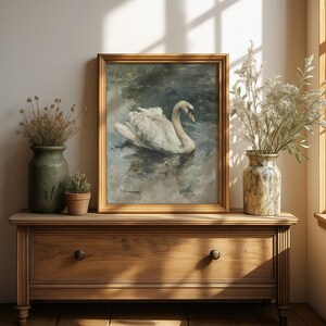 Puede incluir: Pintura enmarcada de un cisne blanco flotando en el agua, en un marco dorado. La obra de arte est&aacute; sobre una c&oacute;moda de madera. Dos jarrones con plantas secas y una peque&ntilde;a planta en maceta completan la escena, creando un ambiente sereno y natural.