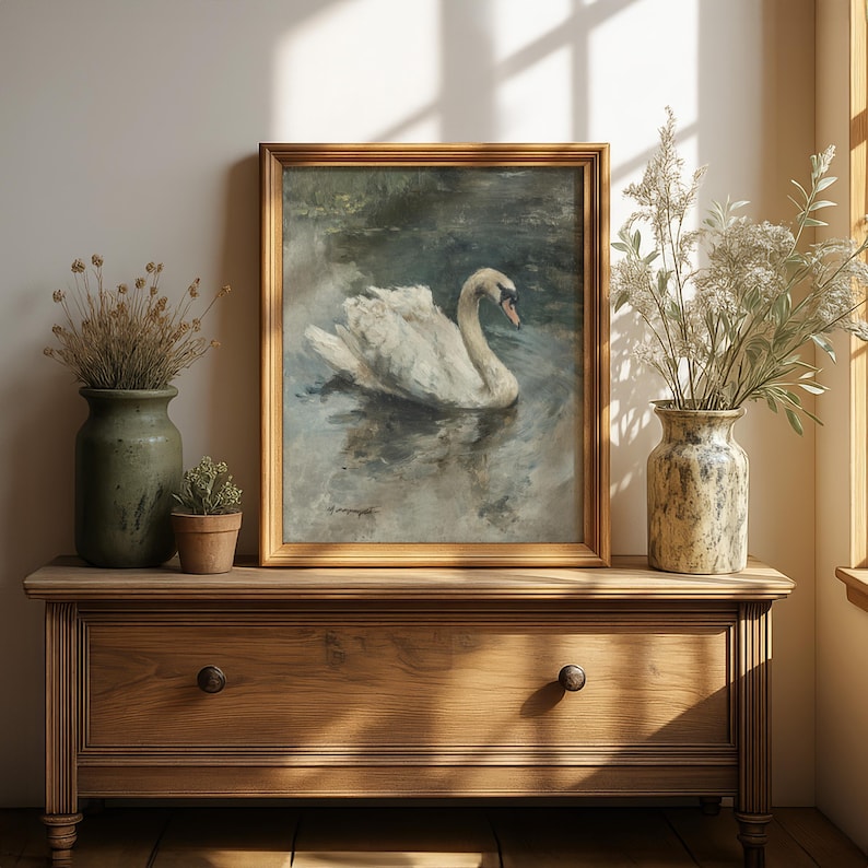 Puede incluir: Pintura enmarcada de un cisne blanco flotando en el agua, en un marco dorado. La obra de arte est&aacute; sobre una c&oacute;moda de madera. Dos jarrones con plantas secas y una peque&ntilde;a planta en maceta completan la escena, creando un ambiente sereno y natural.