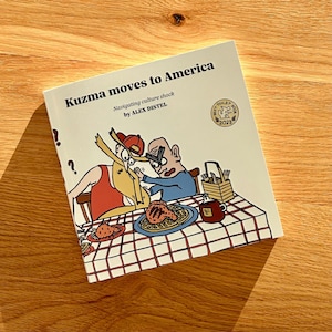 May include: A book titled "Kuzma moves to America" with cartoon illustrations. The cover features a humorous scene of two characters at a table with waffles and a coffee mug. The book is on a wooden surface.