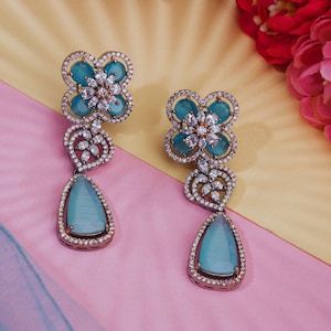 May include: A pair of dangle earrings featuring a blue flower design. The earrings are made of metal and have clear rhinestones. The flowers have a teardrop-shaped blue stone in the centre.
