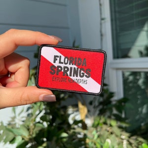 May include: A red and white scuba diving flag patch with black text that reads "Florida Springs Explore New Depths".