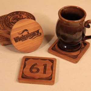 May include: Wooden coasters with engraved designs, including a round coaster with "Vicksburg Mississippi" text, a square coaster with the number "61", and a stack of coasters with state seals. A brown mug sits on a matching coaster.