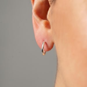 Könnte beinhalten: Eine weiße Keramiktasse mit schwarzem Henkel und dem Text "But First, Coffee" in fetter, schwarzer Schrift. Die Tasse steht vor einem verschwommenen Hintergrund, der eine gemütliche Umgebung suggeriert.