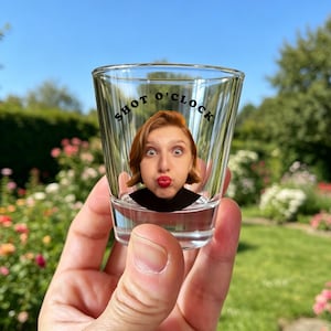 Puede incluir: Un vaso de chupito de cristal transparente con la imagen impresa de una persona con cabello castaño rojizo y labios rojos haciendo un gesto de beso. El texto SHOT O'CLOCK está impreso en letras mayúsculas negras en arco sobre la cabeza.