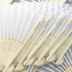 May include: A collection of ivory folding fans with a delicate, openwork design. The fan handles are light wood, and some are inscribed with the text "Jenny & David" in elegant script. The fans are arranged in a layered pattern.