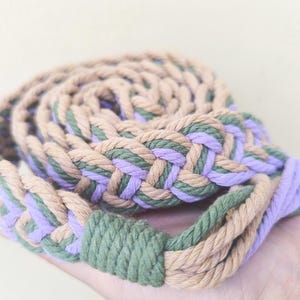 May include: Braided belt in tan, green, and lavender. The belt is made of thick, woven rope, with a braided design. The belt is rolled up, showing the intricate pattern and color combination. A stylish accessory for any outfit.