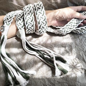 May include: A white and green braided rope belt with silver metal accents and tassels. The belt is tied around a wrist.