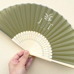 Peut inclure: Éventail pliable vert olive avec un manche blanc. L'éventail porte les initiales "J T" et la date "02.09.2026" imprimées en blanc. L'éventail est ouvert, révélant son design plissé.