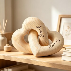 May include: A light-coloured wooden sculpture consisting of two interlocking heart shapes. The wood features a visible natural grain pattern. The names Jack and Emily are engraved in a dark cursive script on the surface of the hearts.