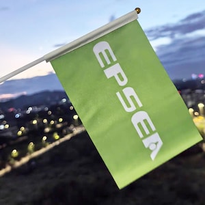 Puede incluir: Una pequeña bandera verde con la palabra "EPSEA" en letras blancas. La bandera está sujeta a un mástil blanco con un remate dorado. El fondo muestra un paisaje urbano borroso al anochecer.
