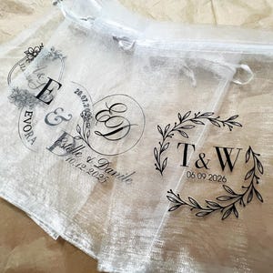 Op de afbeelding: Een set doorzichtige witte organza zakjes met trekkoord en zwarte opdruk. De ontwerpen bevatten bloemenkransen, de initialen E & D, T & W, de namen Estelle & Danile, Evora en de datums 10.12.2025 en 06.09.2026.
