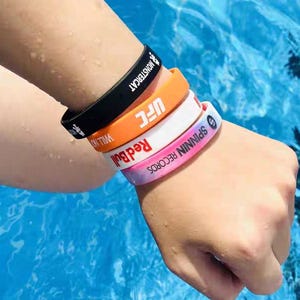 Könnte beinhalten: Fünf bunte Silikon-Armbänder in Schwarz, Orange, Weiß, Rosa und einem Rosa-Blau-Verlauf. Jedes Armband trägt weiße Schriftzüge von Marken und Organisationen. Die Armbänder sind auf einem Handgelenk gestapelt.