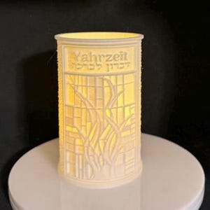May include: A cylindrical, off-white candle holder with the word "Yahrzeit" and Hebrew text. The design features a stained-glass style with a floral motif. The candle holder is illuminated from within, creating a warm glow.