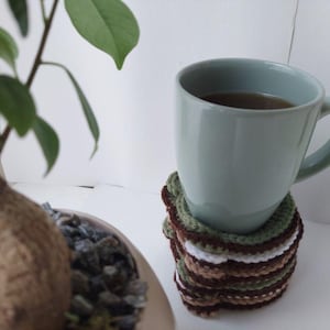 May include: A light blue mug filled with a dark liquid, resting on a stack of crocheted coasters. The coasters feature a mix of brown, green, and white yarn. A small bonsai tree is visible in the background.