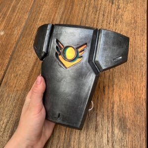 May include: A dark grey, angular chest plate with a stylized emblem featuring orange and yellow accents. The chest plate has a metallic finish and a ring on the side. The item is displayed on a wooden surface.