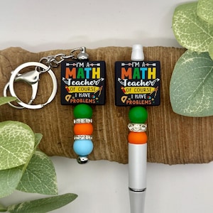 May include: A keychain and a pen featuring a black square with the text "I'M A MATH TEACHER OF COURSE I HAVE PROBLEMS" in colorful letters. The keychain has a silver ring and a beaded design with green, orange, and blue beads. The pen has a silver barrel and a similar beaded design.
