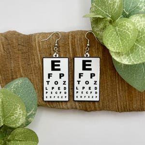 May include: A pair of novelty earrings featuring a white background with black text resembling an eye chart. The letters E, F, P, TOZ, LPED, PECFD, and EDFCZP are displayed. The earrings have silver-colored fish hook ear wires.