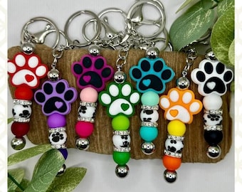 May include: A set of seven colorful paw print keychains with silver key rings. Each keychain features a paw print charm with a small heart in the center, surrounded by colorful beads and silver beads with black and white paw prints.