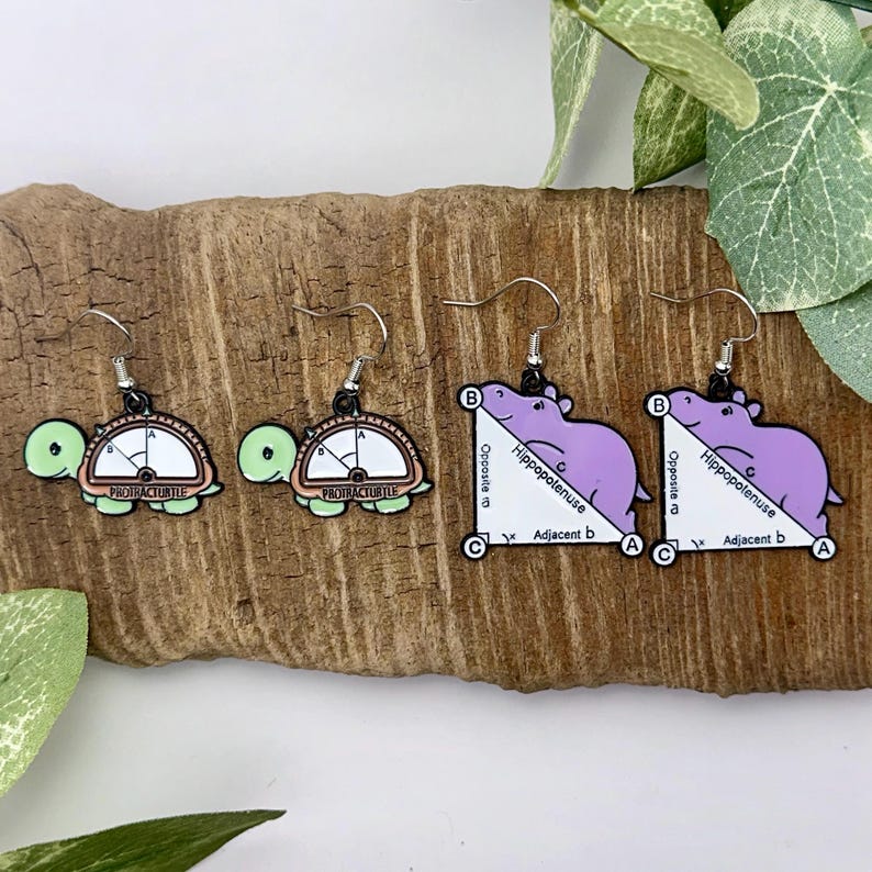 May include: A pair of earrings featuring a green turtle with a protractor design and a pair of purple hippo earrings with a right triangle design. The earrings are on a wooden surface.