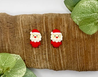 May include: A pair of stud earrings featuring Santa Claus faces with red hats, white beards, and pink skin tones. The Santa figures are crafted with a smooth finish and are positioned side by side, displaying detailed facial features including eyes and noses.