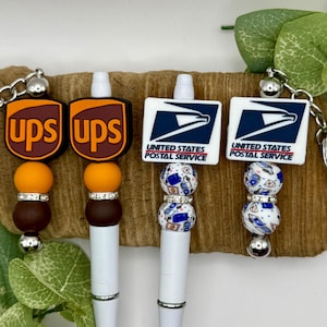 May include: Three white pens with different charms. The charms are a brown and orange UPS logo, a white and blue USPS logo, and a white and blue USPS logo with a white and blue patterned bead.