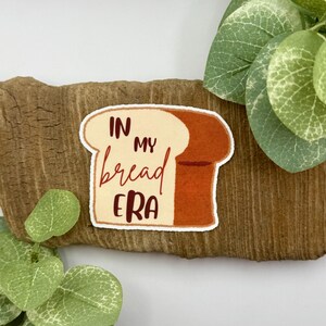 May include: A sticker featuring a slice of bread with the words "IN MY bread ERA" in a handwritten style. The bread slice is beige and brown, with a white border. The sticker is placed on a piece of wood, with green leaves in the background.