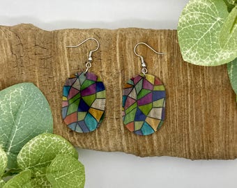 May include: A pair of oval-shaped earrings with a stained glass design. The earrings are made of a clear material with a colorful geometric pattern. The earrings are hanging from silver hooks.