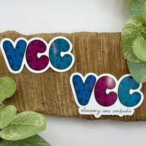May include: Two stickers with the letters "VCC" in blue and purple, filled with paw prints. One sticker has the text "veterinary care coordinator" below the letters. The stickers are on a piece of wood.