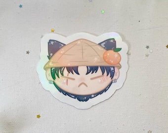 May include: A sticker featuring a cartoon character with a cat-like shape. The character has dark hair, a straw hat, and an orange fruit. The sticker has a holographic effect.