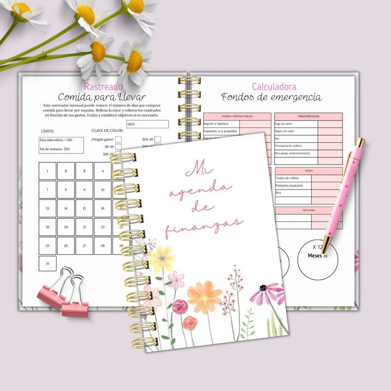 May include: A spiral-bound finance planner with a floral design and the text "Mi agenda de finanzas." The planner includes sections for tracking expenses, with a pink pen resting on the open pages. Two pink binder clips are also visible.