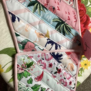 May include: Two quilted pot holders with a pink border. The pot holders feature diagonal stripes of fabric with flamingo, palm tree, and floral patterns in pink, white, and green. One pot holder has the word "Love" printed on it.