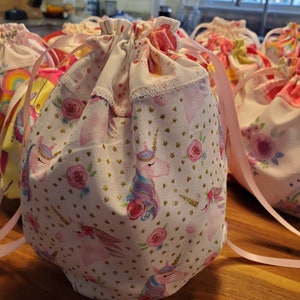 May include: Several drawstring bags with a white background, featuring a pattern of unicorns, pink roses, and gold stars. The bags have pink ribbon drawstrings and are made of fabric. The bags are on a wooden surface.
