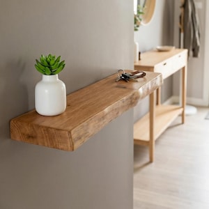 Puede incluir: Estante flotante de madera natural con borde en bruto, montado en una pared gris. Un pequeño jarrón blanco con una suculenta verde está a la izquierda. Llaves a la derecha. Estante de madera maciza.