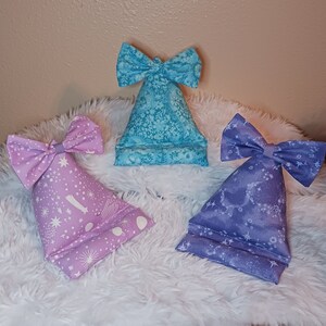 May include: Three fabric phone stands in shades of blue and purple, each with a bow at the top. The stands have various patterns, including floral and celestial designs. The stands are on a white, fluffy surface.