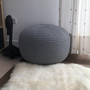 May include: A gray knitted round pouf ottoman sits on a white sheepskin rug.