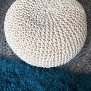 May include: A white knitted round ottoman with a textured surface. The ottoman is sitting on a grey and white rug.